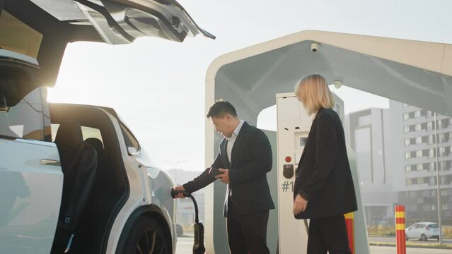 Stylish caucasian woman talking with asian car dealer while buying new electric vehicle. Multiracial people standing near moden auto on charging station. Transport and purchase concept. Electric car.