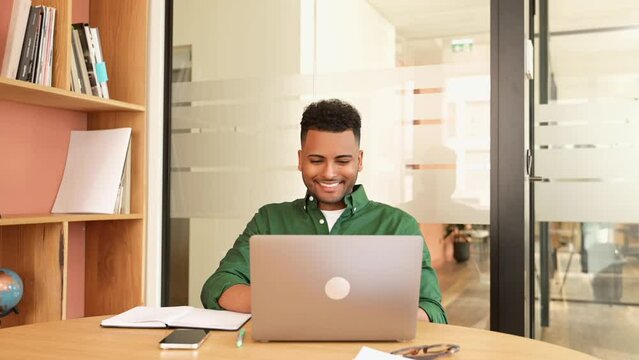 Successful freelancer. Happy millennial Indian man sitting at desk with laptop, smiling and typing, enjoying remote work, male student studying on the distance