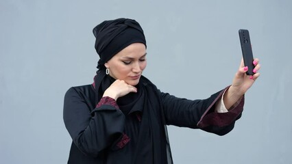 Muslim woman in black hijab makes selfies on modern smartphone. Lady taking selfie enjoys capturing moments on street against grey building