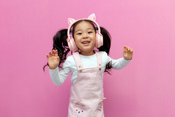 little asian girl in pink children's headphones listens to music and dances on pink isolated...