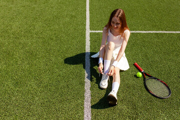 young girl tennis player in white sports uniform ties her shoelaces on sneakers on green court