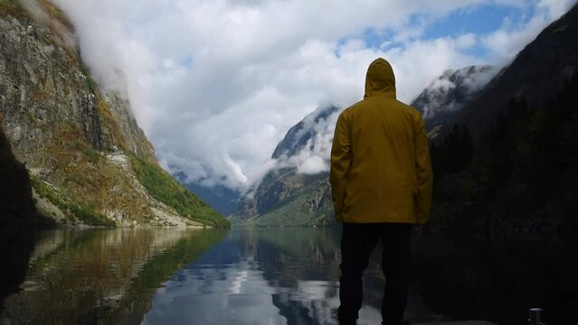 Young man in yellow rain coat enjoying the view of Norways Fjord landscape. impressive mountain reflecting in a calm lake