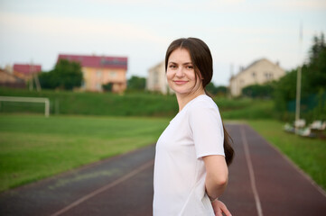 Portrait of young attractive fit girl in black sports tight clothes standing on green field before...