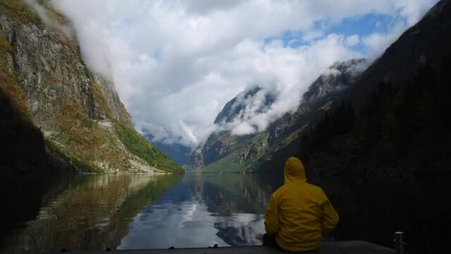 Young man in yellow rain coat enjoying the view of Norways Fjord landscape. impressive mountain reflecting in a calm lake