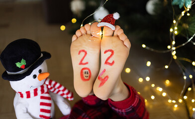 bare feet of a child on which the numbers 2024 are written, luminous garlands, a toy snowman. Cozy,...