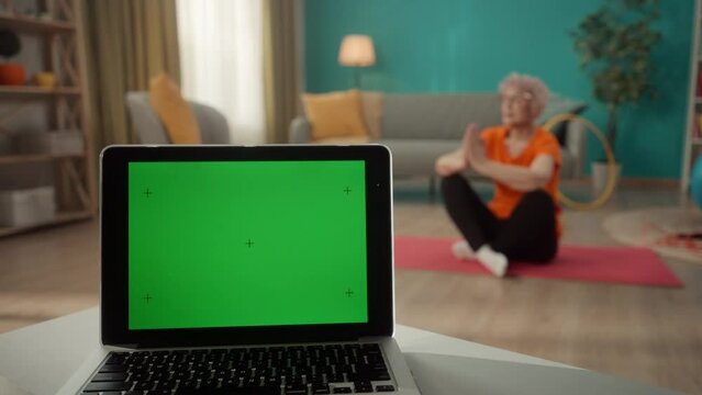 A laptop with a green screen on a table on a blurred background in the living room close up. An elderly woman sits in a lotus position on a sports mat. Advertising area, workspace layout.