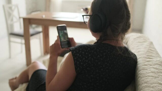 Close-up rear view of girl in black dress and headphones who browses social networks flipping through the phone screen while sitting on the couch at home.