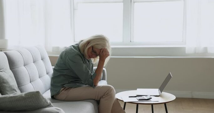Serious blonde elder woman in glasses typing on laptop at home, using calculator for checking domestic payment, finance, getting, frustrated worried bout problems, overspending, money loss,