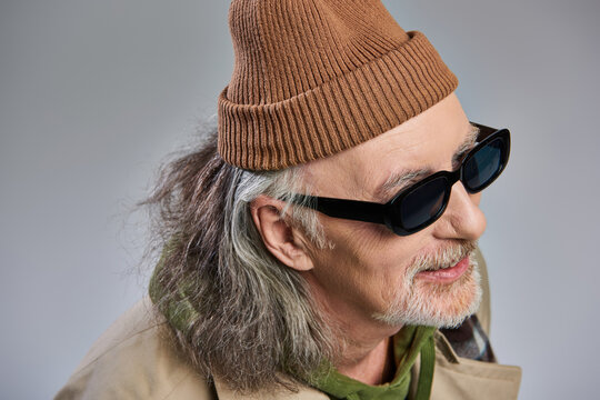 portrait of grey haired and cheerful senior man in fashionable hipster style outfit, beanie hat and dark sunglasses smiling and looking away on grey background, happy aging concept