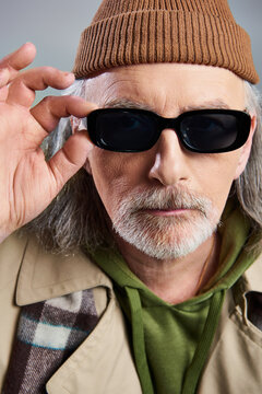 portrait of senior man with groomed beard, in beanie hat and beige trench coat, touching dark sunglasses and looking at camera on grey background, hipster fashion, individuality, aging with style