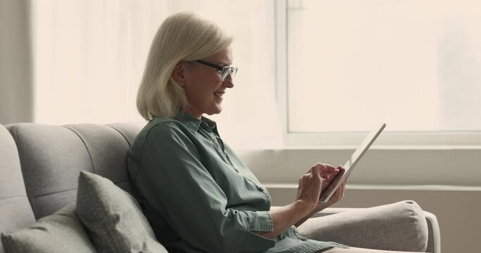 Happy older retired woman in eyeglasses using modern Internet technology at home, holding tablet computer, touching screen, reading, watching online content, looking away, smiling, laughing
