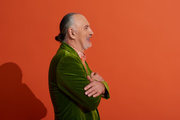 side view of senior male model, optimistic man posing with folded arms and smiling on red orange background, green velour blazer, grey hair and beard, positive and fashionable aging concept