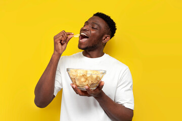 joyful african american man holding plate of chips and biting snack on yellow isolated background