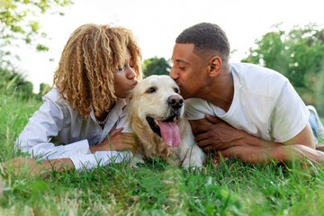 african american happy couple lie together with dog in park in summer, man and woman kiss and love retriever