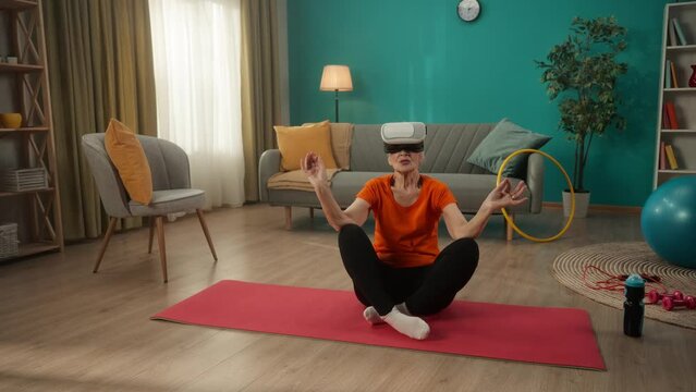 A retired woman in virtual reality glasses sits in a lotus position. Elderly woman doing yoga in virtual reality. Yoga class and technology.