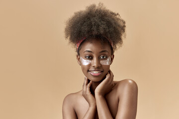 Portrait of beautiful, young, african woman taking care after skin, applying under eye patches against studio background