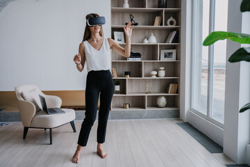 Young fit caucasian woman in white blouse and black pants using vr googles at home, playing game. Pretty Italian student girl educates by virtual reality headset. Remote education and Entertainment.