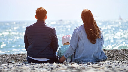 Father and mother with toddler daughter sit on pebble beach illuminated by sunlight. Parents enjoy visiting seaside beach together against shimmering water, backside view © lenblr