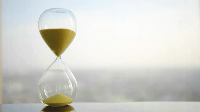 hourglass on table, sand flowing through the bulb of sandglass, time is money concept, times passing concept like work or business deadline, resemble urgency running out of occasion in life