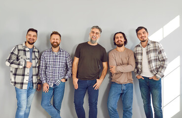 Portrait of a male bearded friends looking cheerful at the camera and smiling on grey wall background. Full length photo of a group of brutal friendly men laughing in casual clothes.