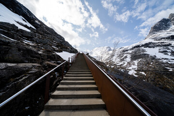 now on the mountain and the rocks are gray but above them is a blue sky with white clouds where the steps of a gray concrete staircase with rusted metal railings lead