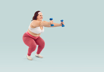 Full length photo of a funny happy young fat overweight woman wearing sportswear doing exercise with dumbbells isolated on studio blue background. Workout sport, fitness and body positive concept.