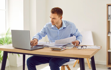 Fototapeta premium Serious, busy accountant working on computer in office. Young man sitting at his desk, using laptop, doing business paperwork, working with financial documents