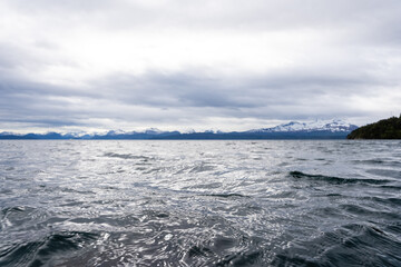 Norwegian fjord with blue water and overcast sky, but in the distance you can see a straight horizon above which dark mountains with white snow open