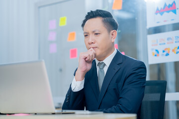 Smiling middle-aged executive asian businessman sitting at a worktable a modern office, typing on computer keyboard, and talk to work, sending emails to his business partners, working on marketing.