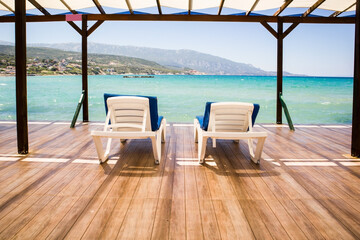 White sun loungers with blue cushions are waiting for their guests on the pier by the sea.