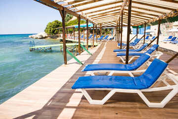 White sun loungers with blue cushions are waiting for their guests on the pier by the sea.