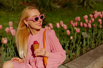 Happy smiling fashionable blonde woman wearing trendy pink sunglasses, pleated dress, holding fruit ice cream, posing in street, among flowers. Copy, empty space for text