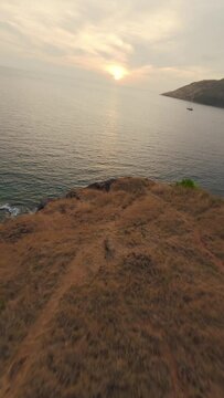 Vertical video 4k. Aerial view dive from mountain rock sea shore flying over water surface yacht boat tropical island at sunset. FPV sport drone low shot exotic beach lagoon ripple calm pond surface