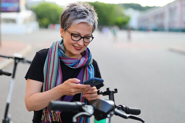 Smiling mature woman using smartphone to rent an electric scooter in the city.