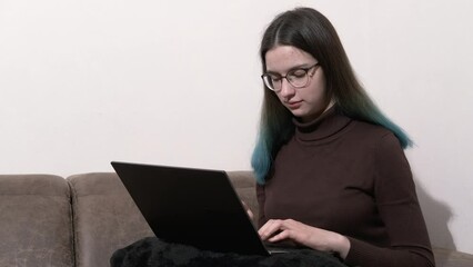 An emotional teenage girl with blue hair using a laptop, sitting at home on the couch, typing, shopping or chatting online on a social network, working on a project, an exam. Online education.