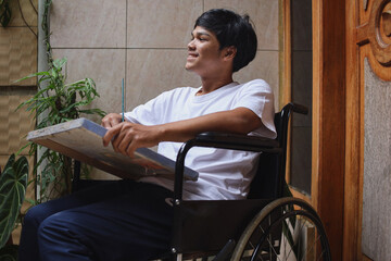 Disabled Asian man on wheelchair smiling and looking for inspiration for his painting art work at home