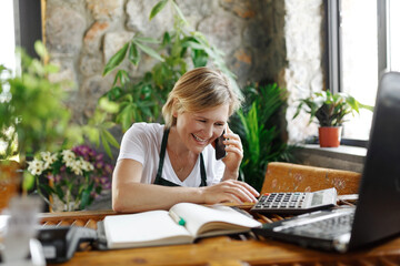 Young woman entrepreneur sitting at desk and typing on computer while chatting on smartphone. Happy female florist in green floral center tapping on laptop and counts on calculator - obrazy, fototapety, plakaty
