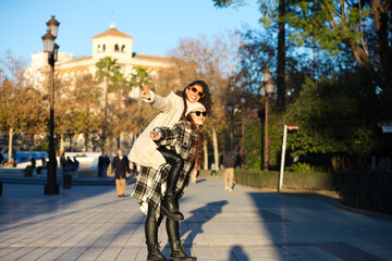 Obraz premium two young, beautiful Spanish women who are friends have fun playing on top of each other while they are walking and enjoying the sunny winter day. The women are wearing winter clothes.