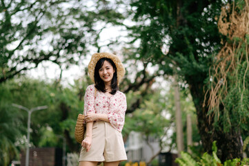 Naklejka premium Portrait of asian young woman traveler with weaving hat and basket happy smile on green public park nature background. Journey trip lifestyle, world travel explorer or Asia summer tourism concept.