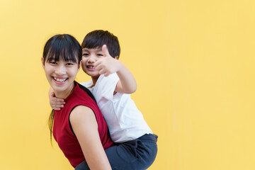 Happy cheerful Asian woman and a little young boy playing together, a woman piggyback or carrying a little boy on her back. Woman and boy portrait on yellow pastel background.