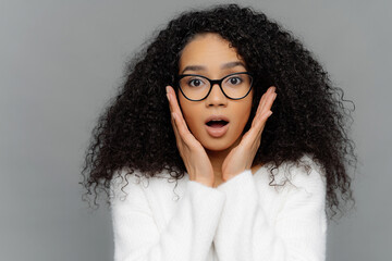 Surprised dark-skinned woman, curly hair, touches cheeks, shocked expression, white jumper, grey background