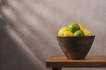 Wooden bowl with lemons and limes on wooden bench against concrete wall with shadow. Cozy home interior in minimalist style