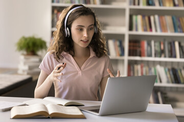 Focused pretty student girl in headphones talking on video call at laptop in university library, sitting at computer, open books, consulting online teacher, attending learning Internet conference