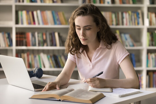 Serious Focused Pretty Student Girl Studying In Campus Library, Reading Book, Textbook At Table, Writing Notes, Working On Essay, Article, Research At Tdesk With Laptop Computer