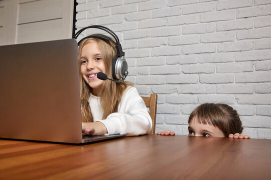 Little girl watches her older sister's online class. In a room with a white brick wall, a teenager sits at a table in a headset with a laptop, next to it is a small child watching