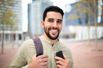 Smiling young middle eastern man holding mobile phone walking outside in the city