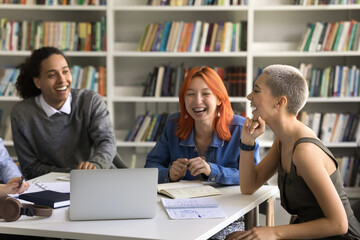 Cheerful funky diverse college friends talking and laughing in campus library, having fun. Classmates doing homework together, sitting at table with laptop and notebooks