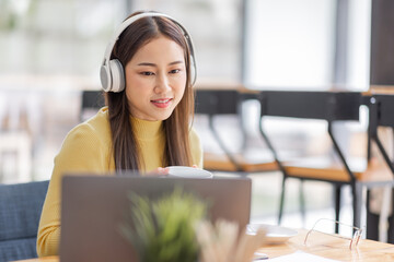 Young adult happy smiling Asian student or business woman wearing headphones talking on online chat meeting using laptop in office or campus, asian female student wear glasses learning remotely.