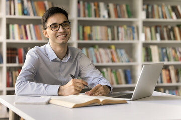 Cheerful smart young student guy in glasses working on essay, research study in campus library, studying at textbook, writing notes at laptop, looking away, thinking, smiling, laughing