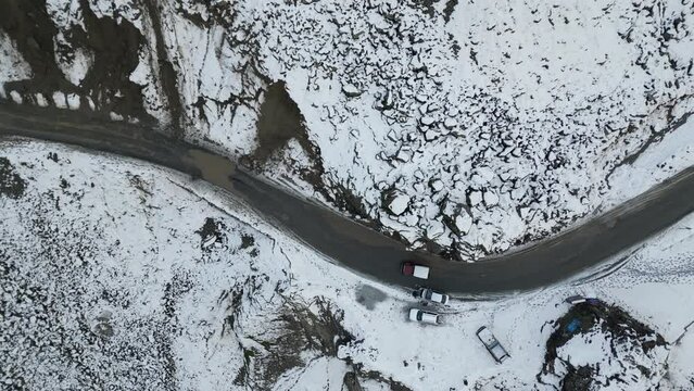 Jeep running on Karakoram highway over a sharp road curve in a snow-capped remote mountainous region near Dasu hydropower project Upper Kohistan. Few vehicles are parked on side of road.